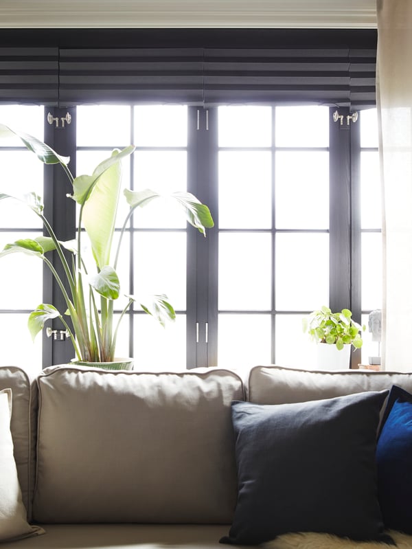 A light beige sofa-bed stands in front of four windows with beige curtains and dark grey pleated block-out blinds.