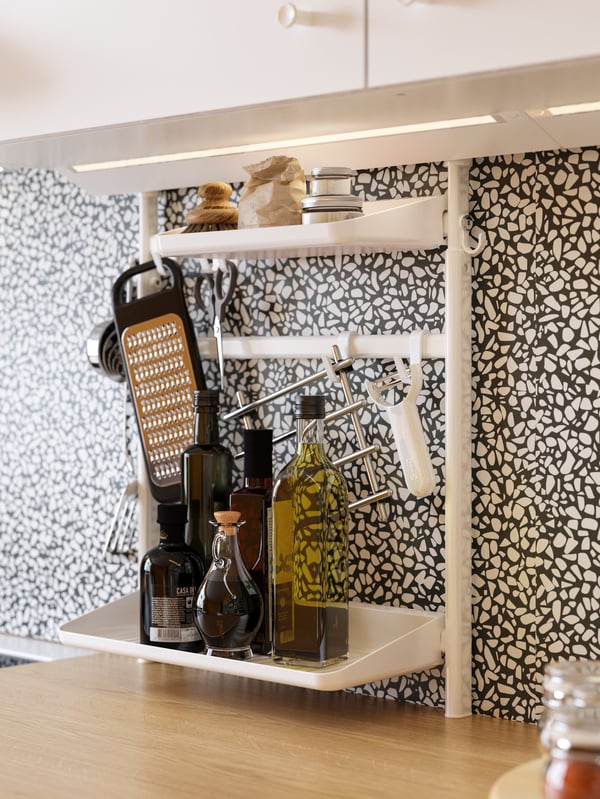A white SUNNERSTA kitchen organiser holding a grater, bottles of condiments and other kitchen items above a worktop.