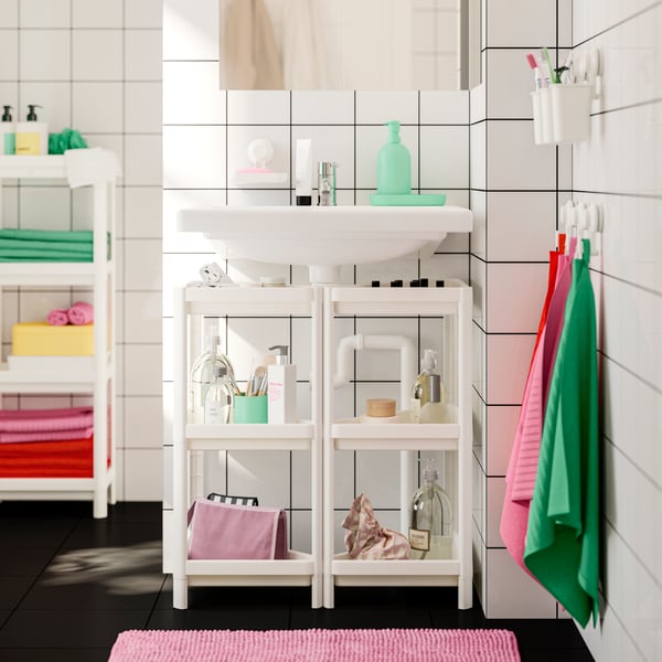 Sous une vasque, deux étagères d’angle VESKEN blanches remplies d’articles de toilette. Des serviettes roses et vertes sont suspendues au mur.