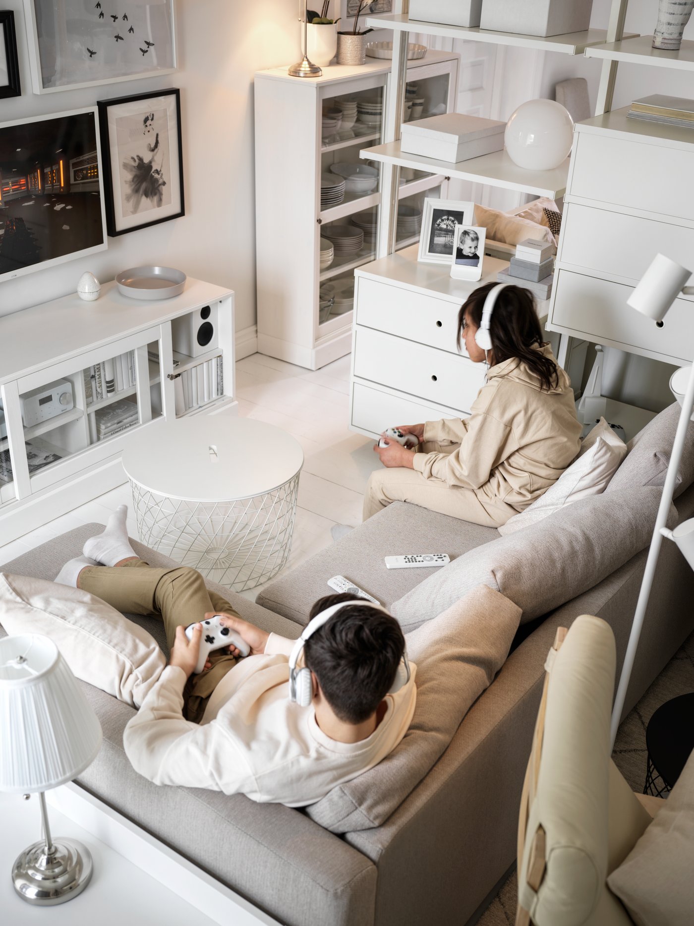 A living room in shades of white, with two teens in a beige FRIHETEN corner sofa-bed playing a video game, consoles in hand.