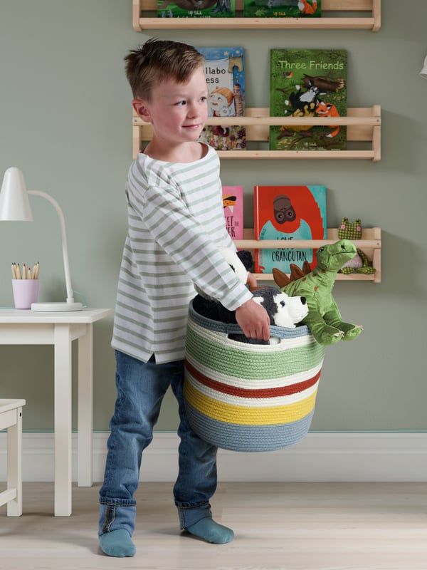A child is carrying a STRANDSKATA basket with plush animals inside. Behind them is a bookshelf with childrens books.