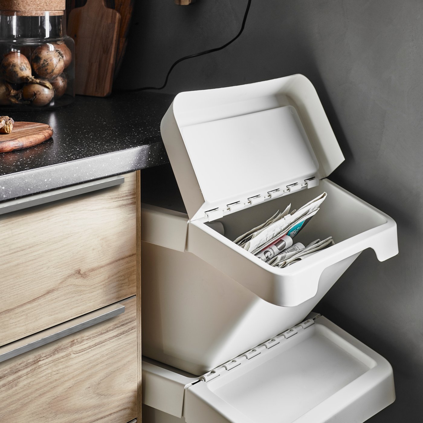 Un bac de tri de déchets SORTERA blanc avec un couvercle ouvert qui laisse voir des journaux à l’intérieur est posé sous une tablette de bocaux en verre de conserves rouges.