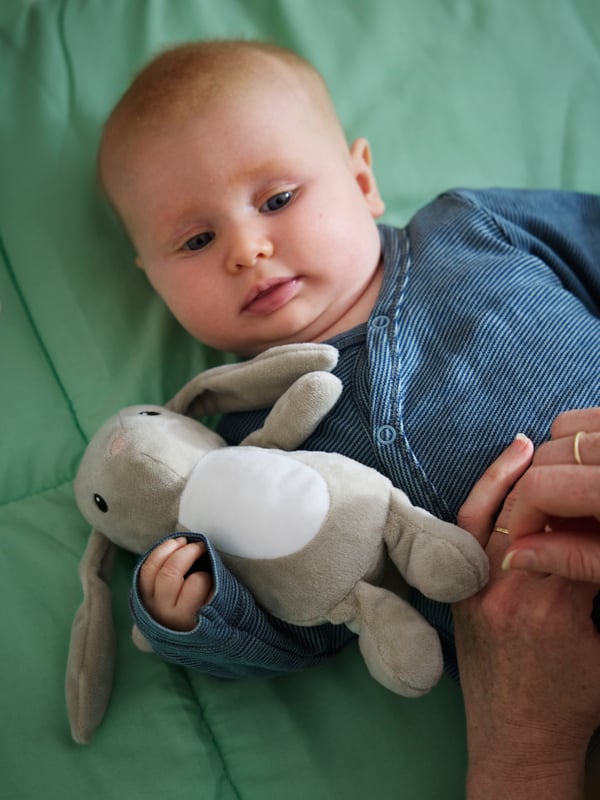 Gros plan sur un bébé en pyjama bleu couché sur le sol et tenant un lapin en peluche DVÄRGHARE.
