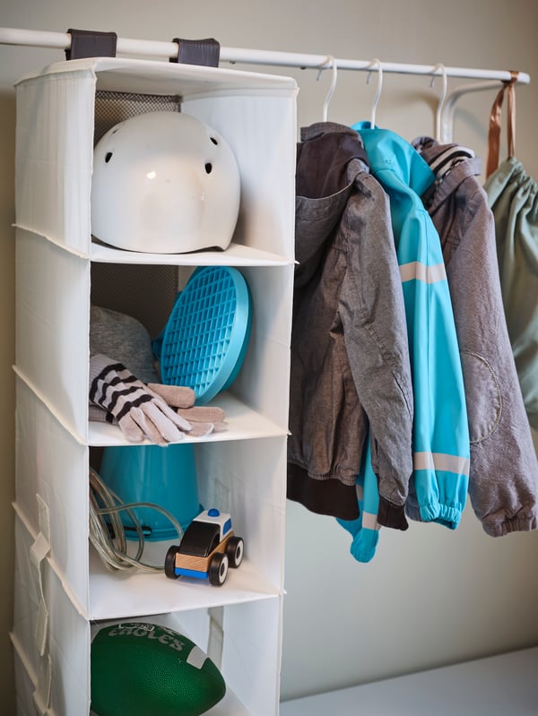 Two MULIG clothes bars hold jackets and a RASSLA storage with 5 compartments containing a helmet, toys and other things.