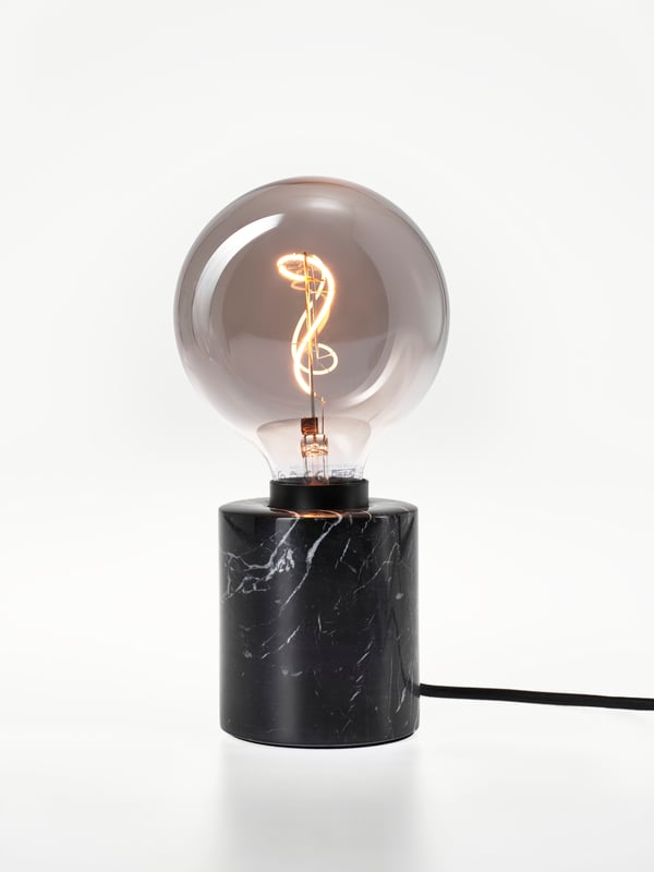 A lit LED bulb with a smokey grey glass globe. It’s installed in a short black base in a white space.