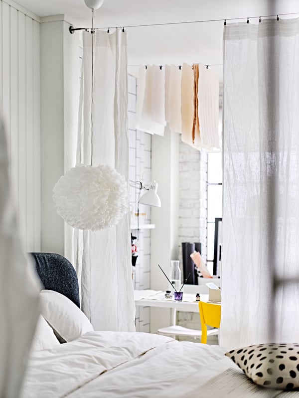 White DYTÅG curtains separating a work area from a sleeping area, with a white VINDKAST pendant lamp above a dressed bed.