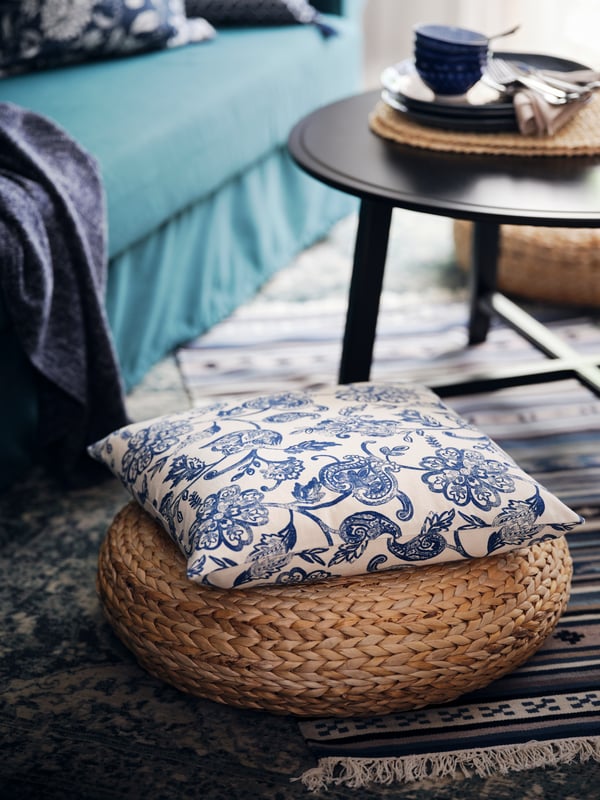 Un coussin revêtu d’une housse JUNIMAGNOLIA posé sur un siège en fibres de bananier ALSEDA, à côté d’une table KRAGSTA noire.