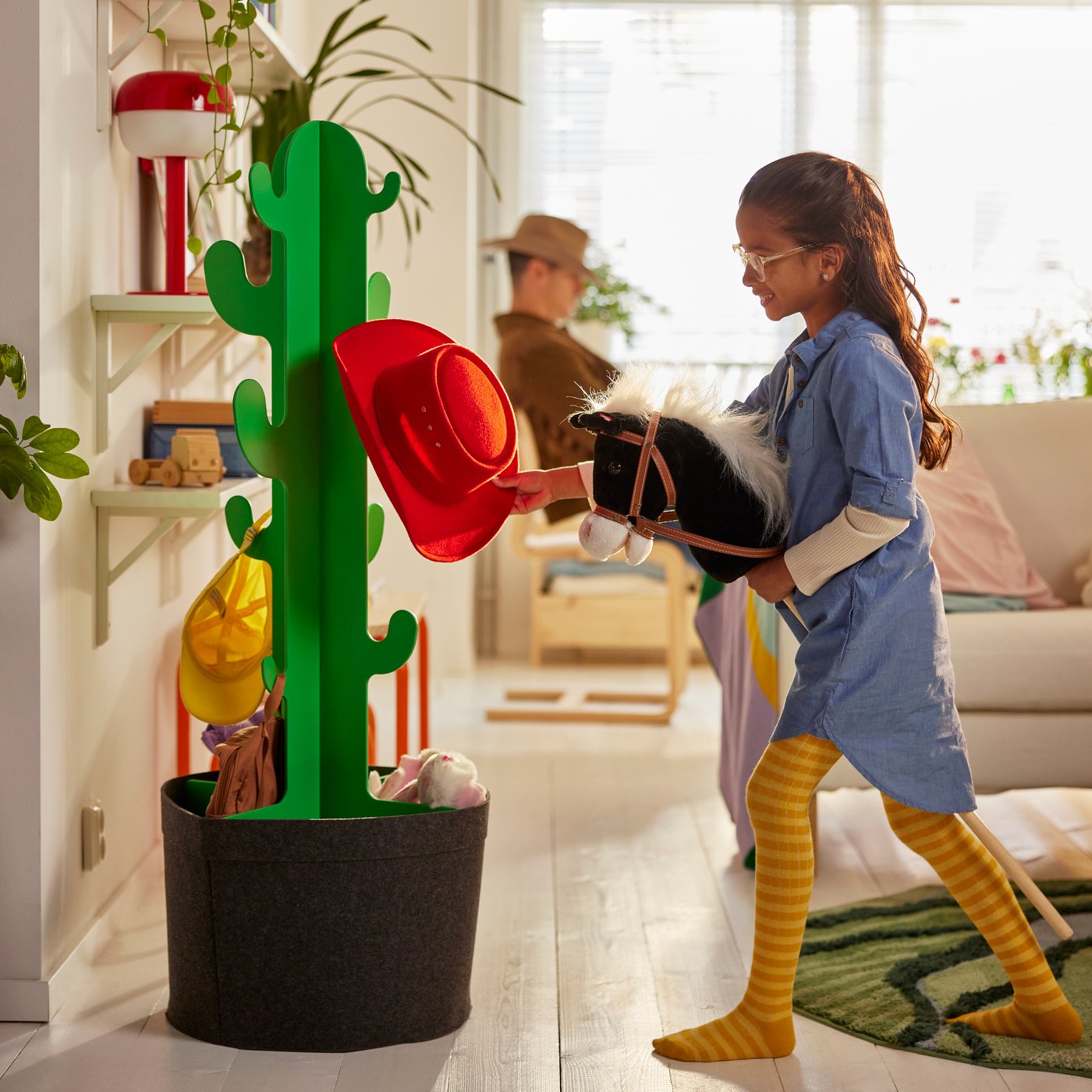 In a sunlit living room, a child on a hobby horse hangs a red cowboy hat on a cactus-shaped GREJSIMOJS clothes stand.