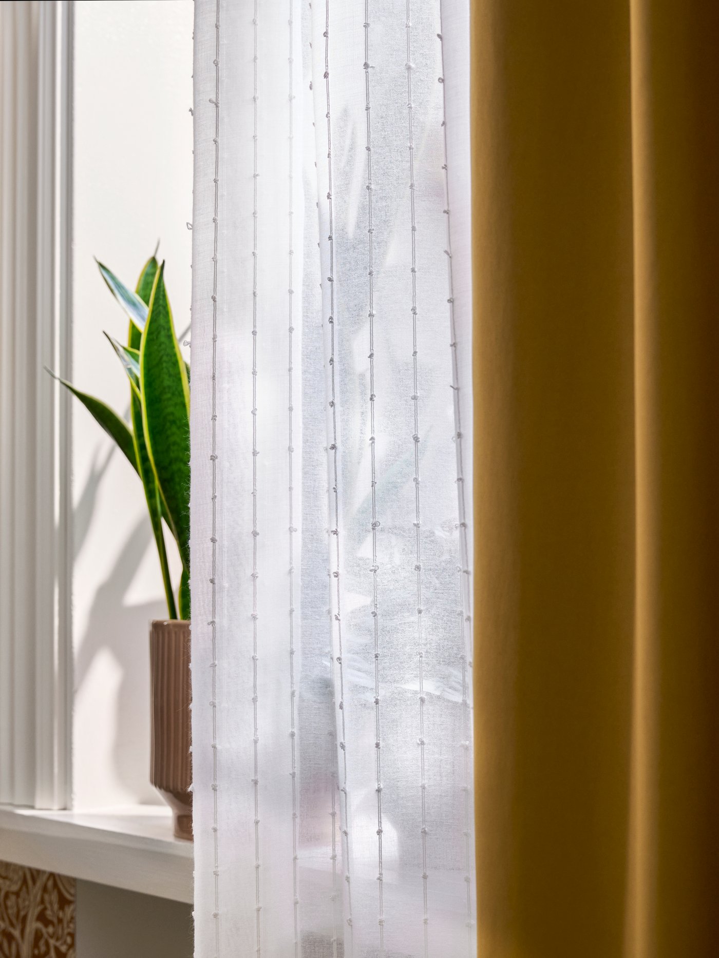 MATILDA transparenter Vorhang in Weiß hängt am Fenster, Sonnenlicht fällt neben gelbem Vorhang und Pflanze hindurch.