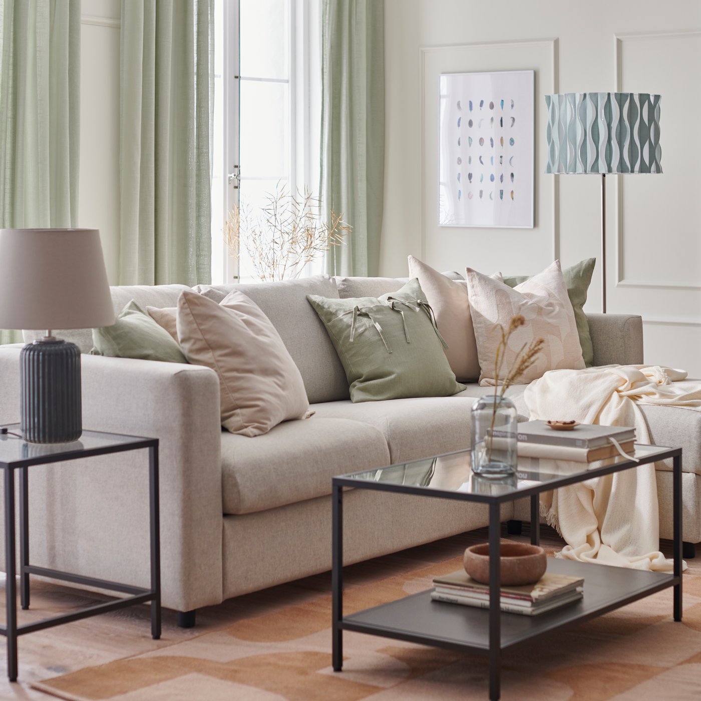 Living con sillón blanco decorado con cojines y mantas en tonos grises y rosados. Además hay cortinas verdes y una mesa de centro de vidrio.