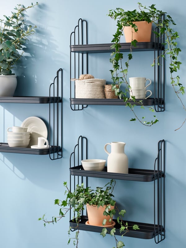 Three black APOTEKARE wall shelves, with two levels, mounted on a light blue wall, holding cups, bowls, and potted plants.