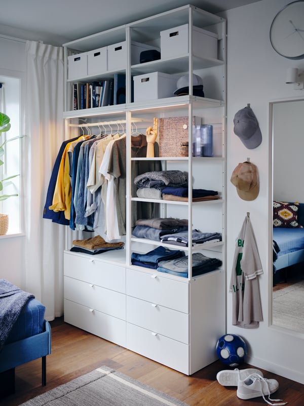 A white PLATSA wardrobe with open and closed storage, integrated lighting, shirts on hangers, folded clothes and TJENA boxes.