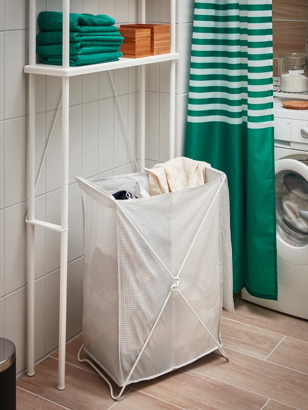 Baño con lavandería integrada. Un canasto de ropa TORKIS con ropa a su interior. Una estructura de DYNAN blanca que contiene toallas de baño DRAGAN.