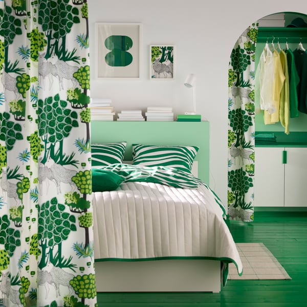 A room with a bed that has green cushions and a white bedspread, behind forest-patterned ÄLGÖRT pre-cut fabric curtains.