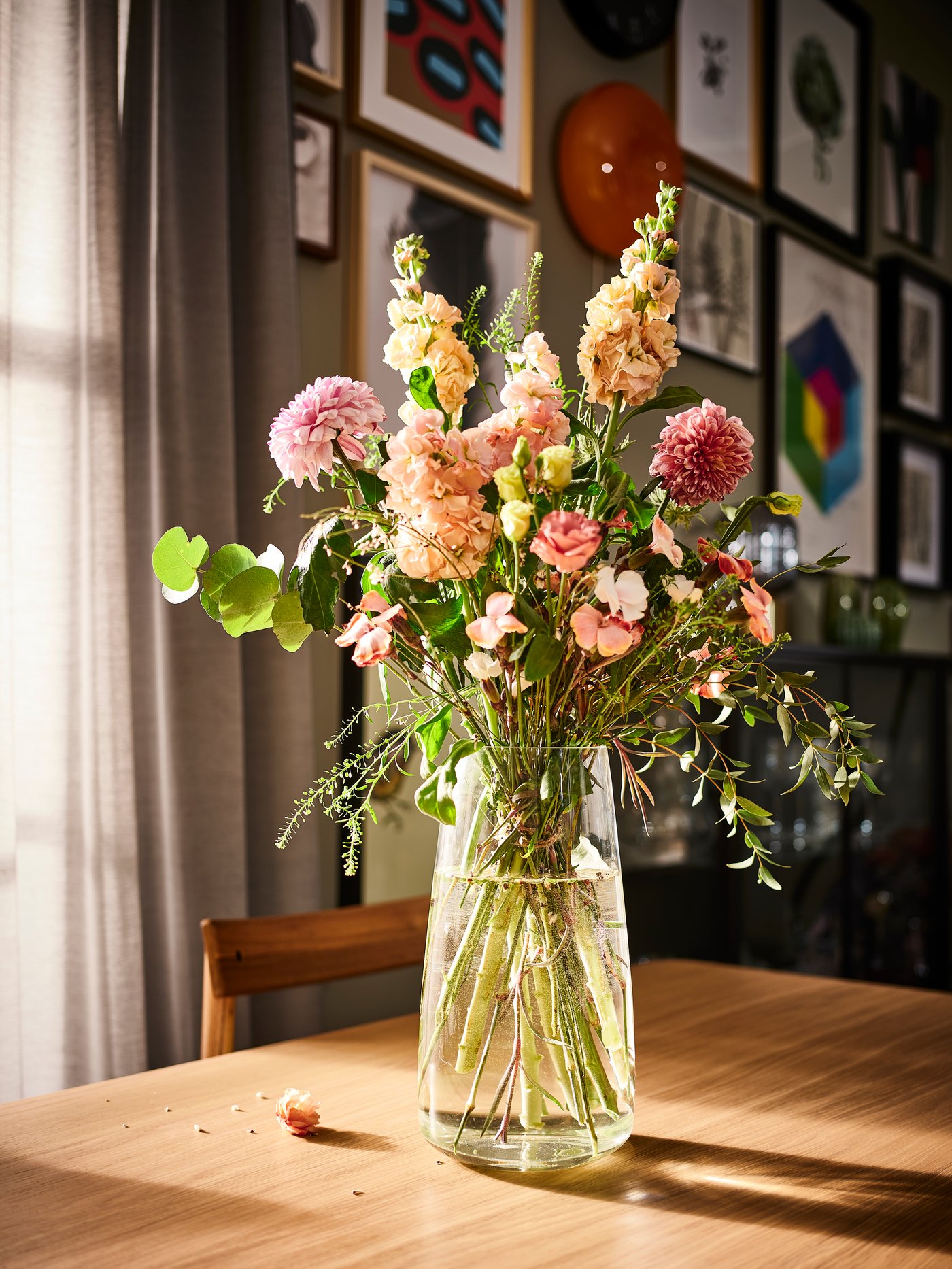 Fresh flowers in a transparent vase sit on a sunlit table.