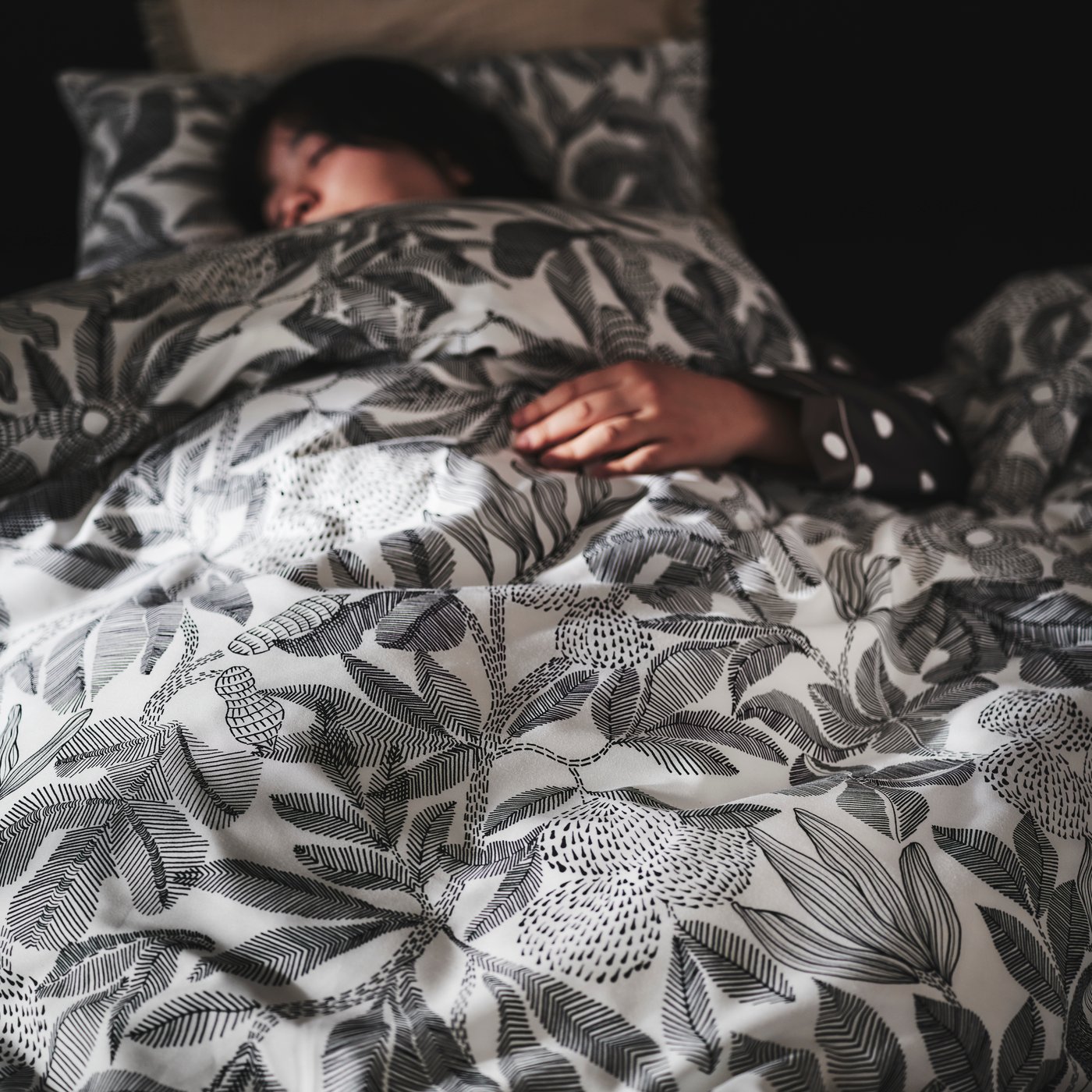 A person asleep in a SLATTUM upholstered bed under a duvet that’s covered in black and white BROKDRACENA bed linen.