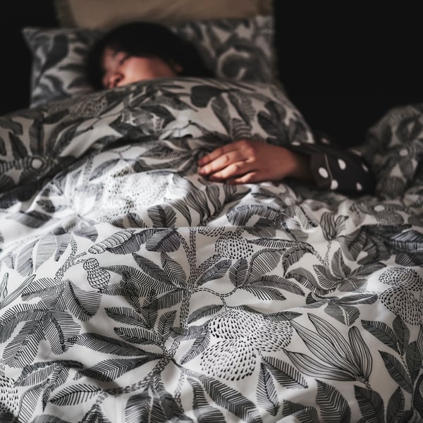 Pessoa a dormir numa cama estofada SLATTUM sob edredão com roupa de cama BROKDRACENA em preto e branco.