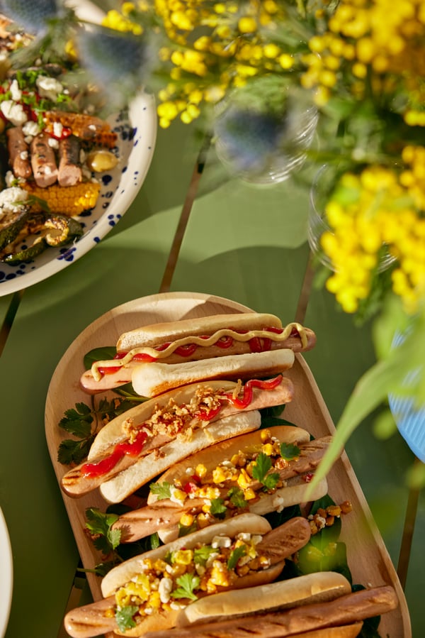 Différents hot-dogs avec des garnitures colorées sont disposés sur un plateau en bois, à côté d'une composition florale sur une table verte.