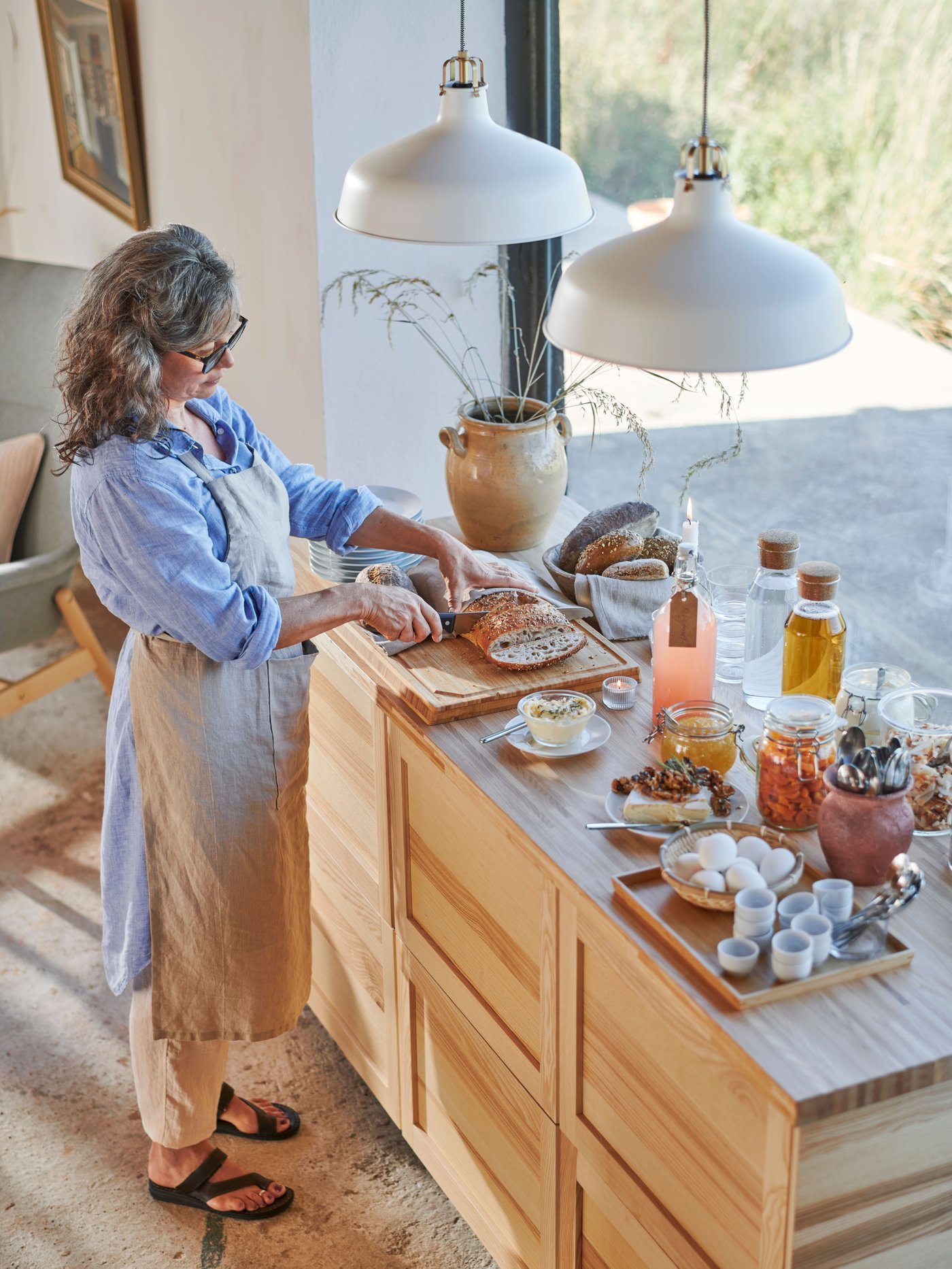 A person, wearing an apron, is standing next to a kitchen island, slicing bread on an APTITLIG butcher’s block.