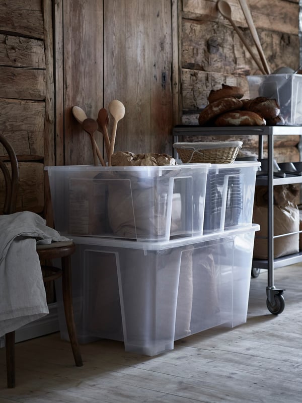 Two SAMLA clear plastic tubs filled with storage on the floor in a garage type setting.
