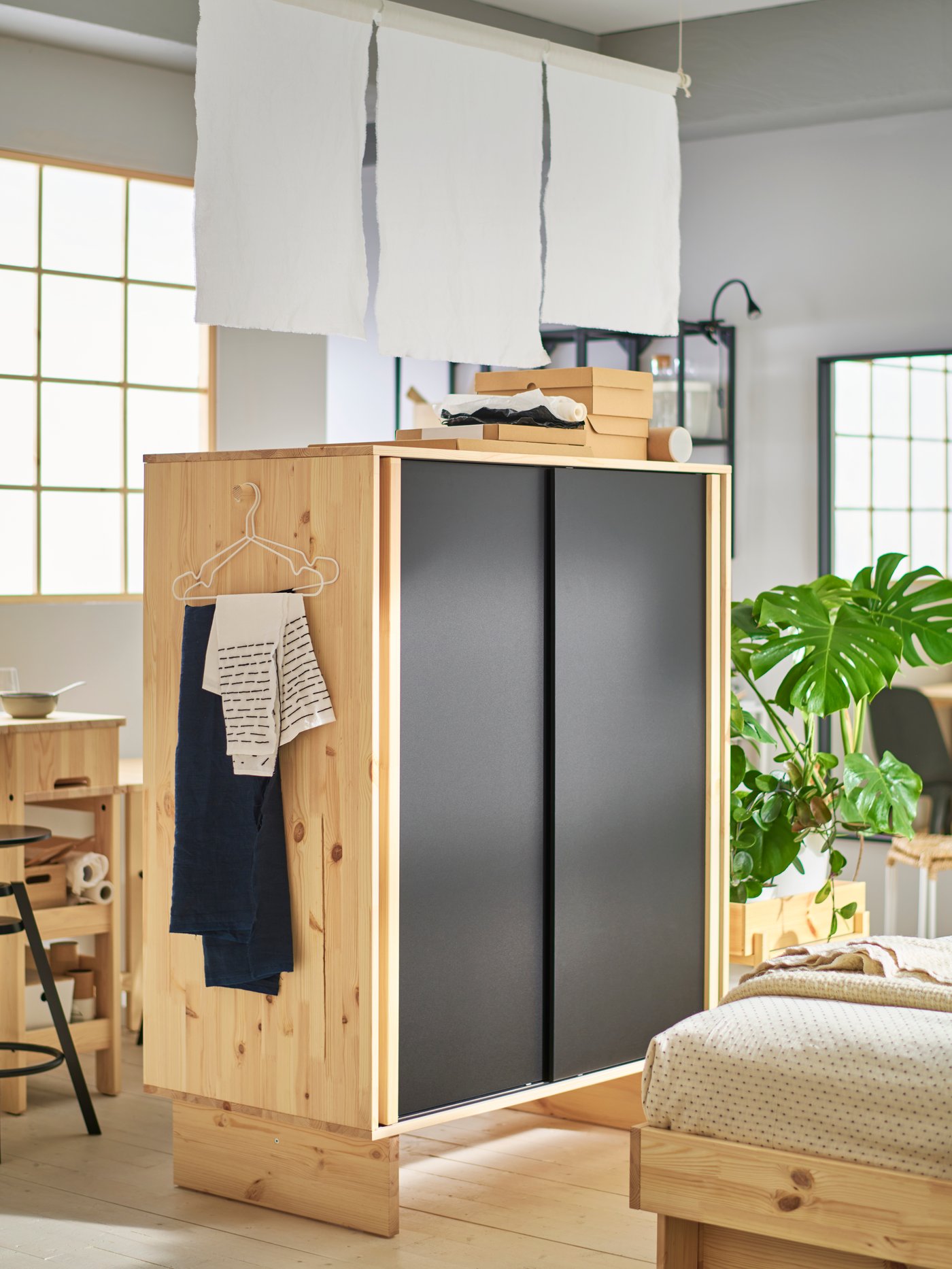 A GLAMBERGET wardrobe made of untreated wood with black sliding doors, placed in the middle of the room as a room divider.