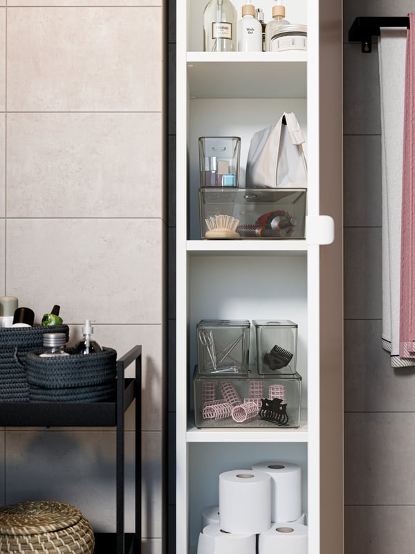 An open high cabinet in white with transparent boxes, toilet paper, lotions and more inside. A black trolley is beside it.