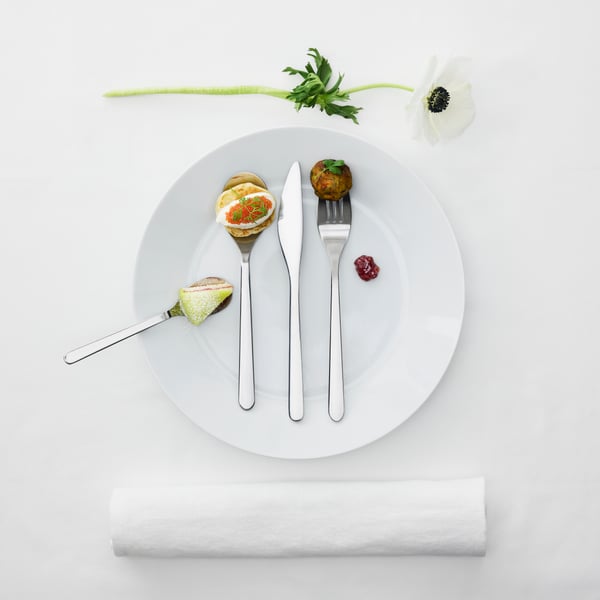 A set of FÖRNUFT utensils on top of a white plate. Each utensil holds a different piece of food, like cake and caviar.
