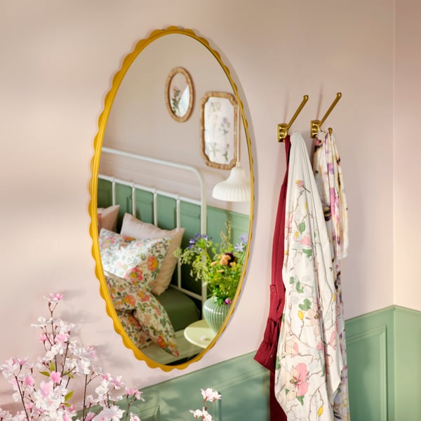 Ein dunkelgelber DAGGKAPRIFOL-Spiegel reflektiert ein gemütliches Schlafzimmer mit Blumendekor und einem Bett, das in sanften Farben gehalten ist.