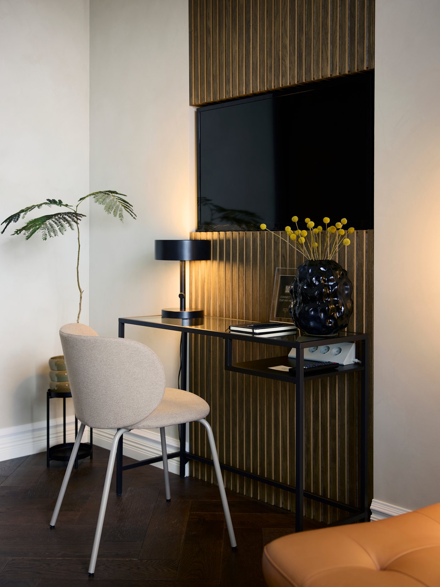 A VITTSJÖ laptop table with a distinct black-brown frame and glass top is in a hotel room below a TV, by a dark beige chair.