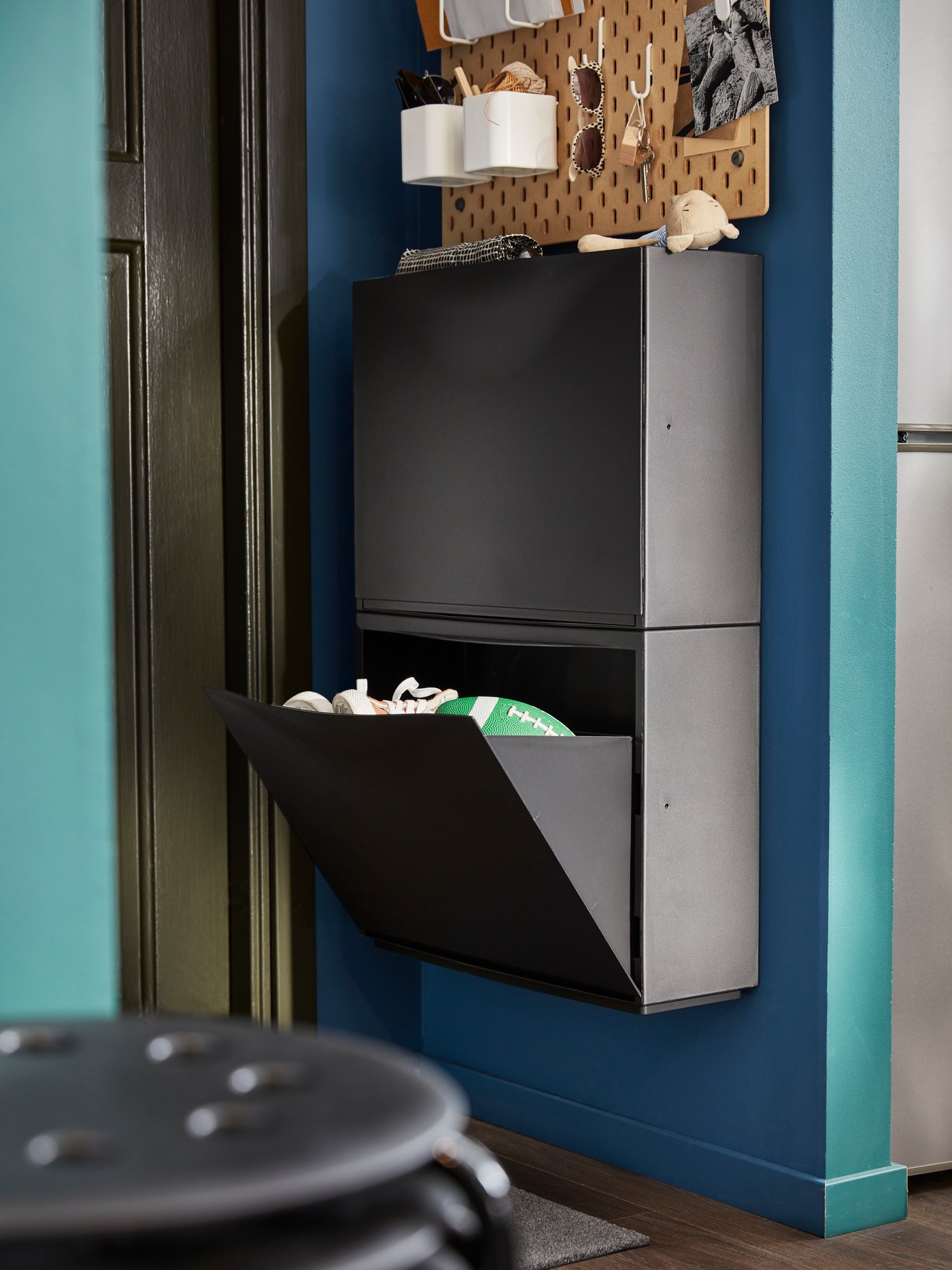 Hallway with blue wall, open shoe cabinet with green football and shoes, wall rack with sunglasses, scissors, and toy.