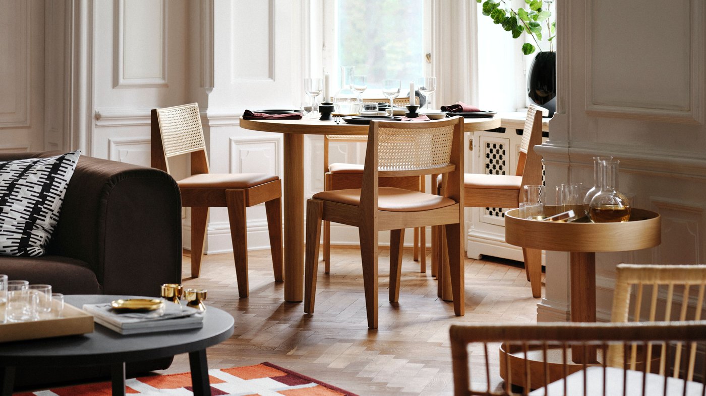 A living room dining set with an oak veneer STOCKHOLM 2025 table and oak and rattan chairs placed by large bay windows.