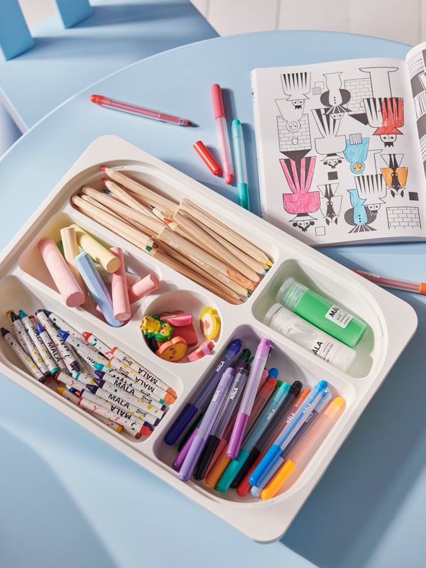 A white TROFAST storage tray with compartments on a light blue table filled with MÅLA felt tip pens and other art materials.