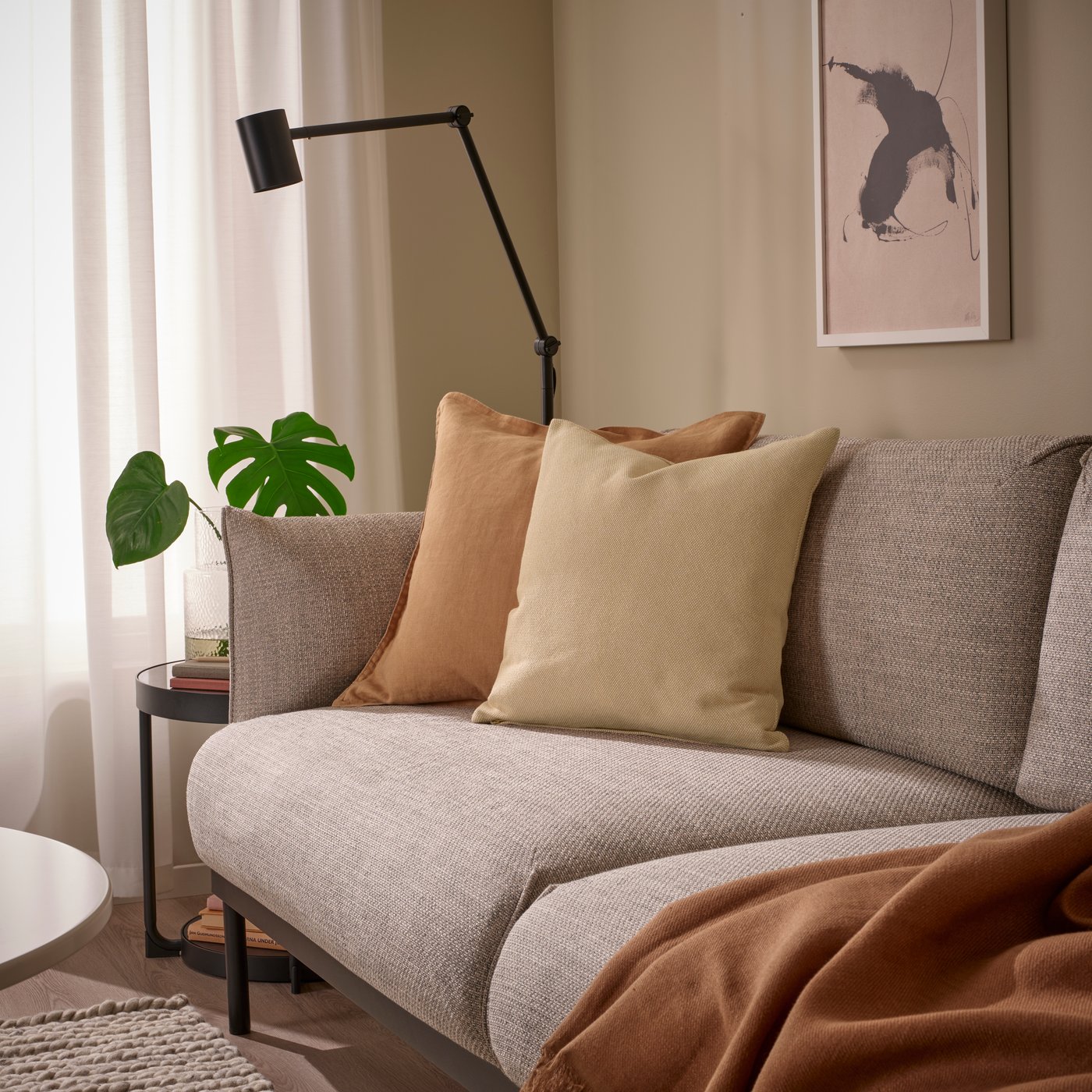 Auf einem hellgrauen Sofa liegen ein gelbbeiges SANDTRAV Kissen aus recyceltem Polyester mit robuster, griffiger Struktur, ein hellbraunes LAGERPOPPEL Kissen und ein passendes, braunes Plaid.