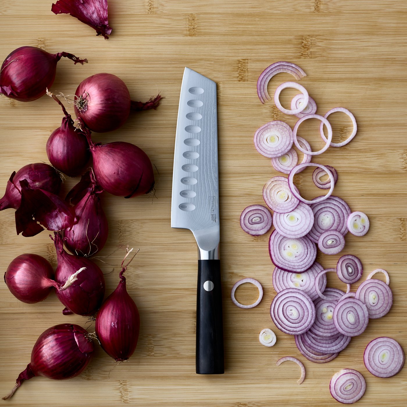 A close-up of a black FJÄLLRÖDING santoku knife made of Damascus steel, next to chopped red onions on a wooden board.