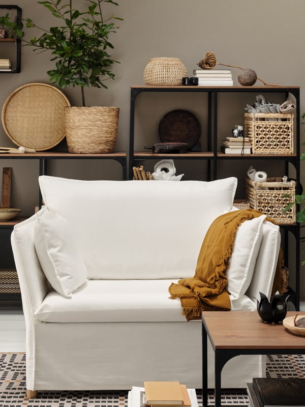 Un fauteuil 1,5 place blanc, un jeté brun doré foncé, des étagères noires avec des paniers, des livres et d’autres objets.