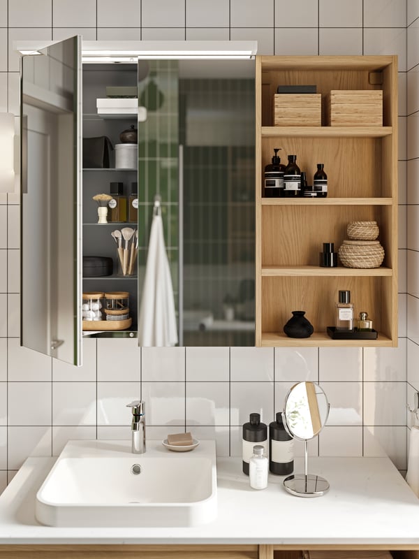 A LETTAN mirror cabinet has one door open and next to it is an open wall cabinet with bathroom products on the shelves.