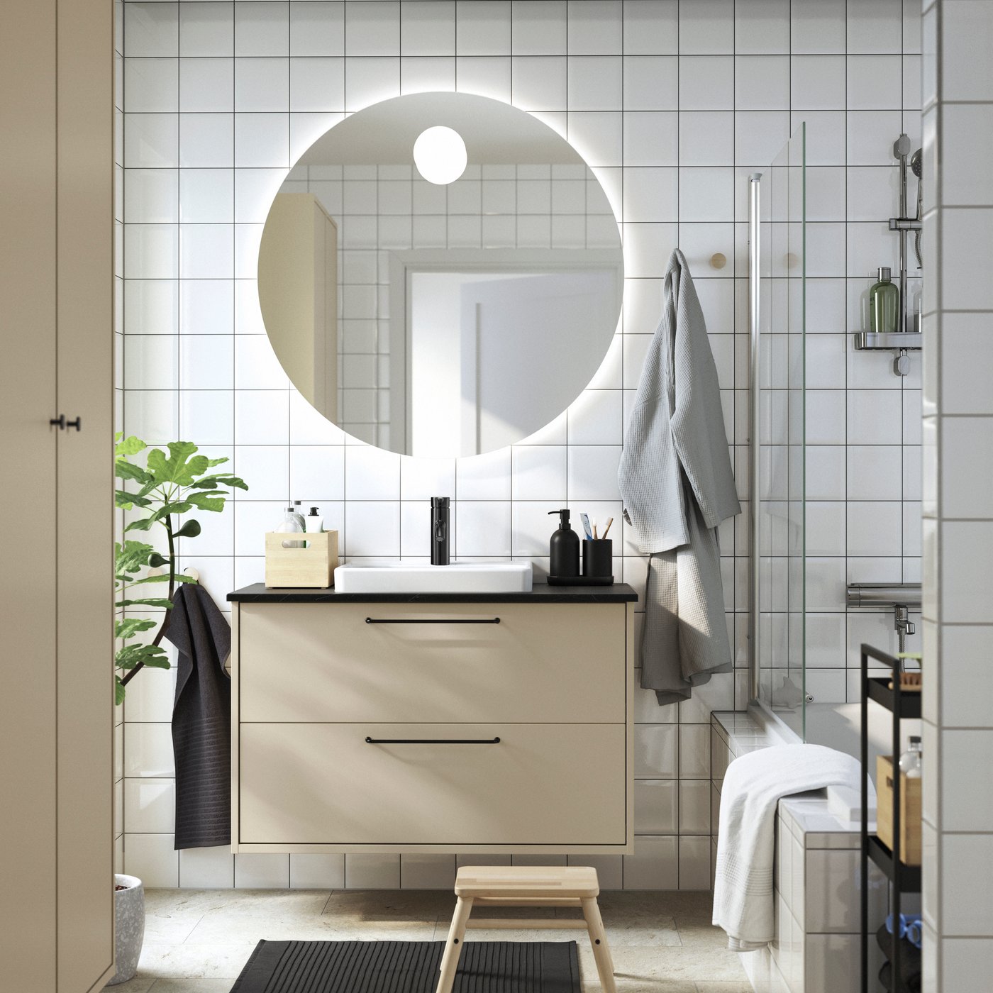 A bathroom with white tiles and a large round mirror, a beige HAVBÄCK wash-stand with black handles.