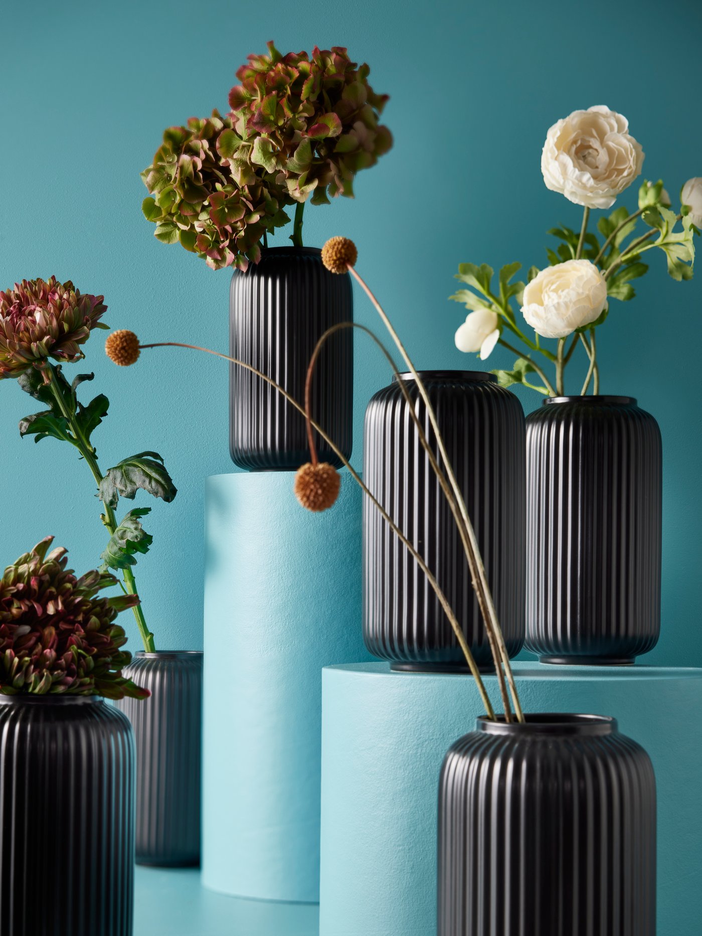 Six black STILREN vases, all holding flowers except one, some placed on pedestals in a light blue space.