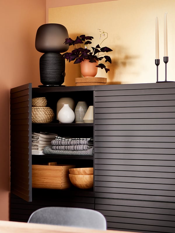A black BESTÅ/STOCKVIKEN cabinet with one door open showing table linen, vases and bowls, with a speaker lamp on top.