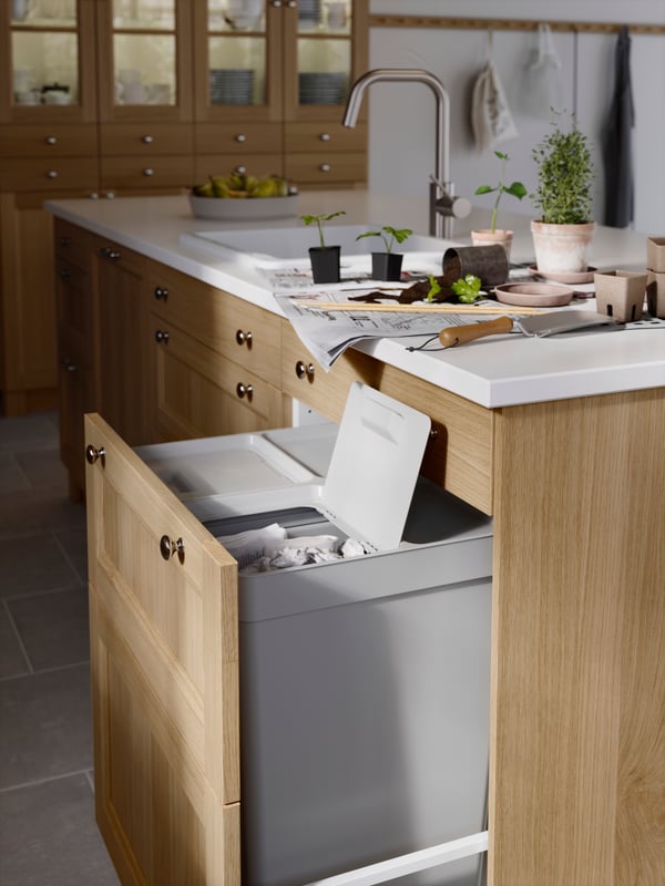A pull-out door in a kitchen with a white marble effect countertop, holding HÅLLBAR waste bins. One of the bin lids is open.