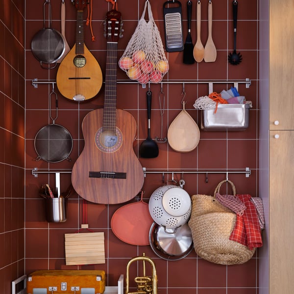 Mur revêtu d’un carrelage brun-rouge et portant toutes sortes d’objets accrochés à des barres supports KUNGSFORS : ustensiles de cuisine, fruits, instruments de musique, etc.