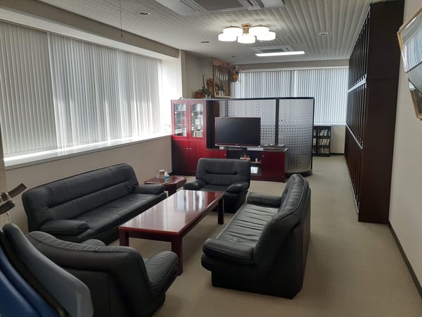 黒いソファとローテーブルを向かい合わせに配置した応接室。テレビと収納棚があり、窓から光が差し込んでいる。