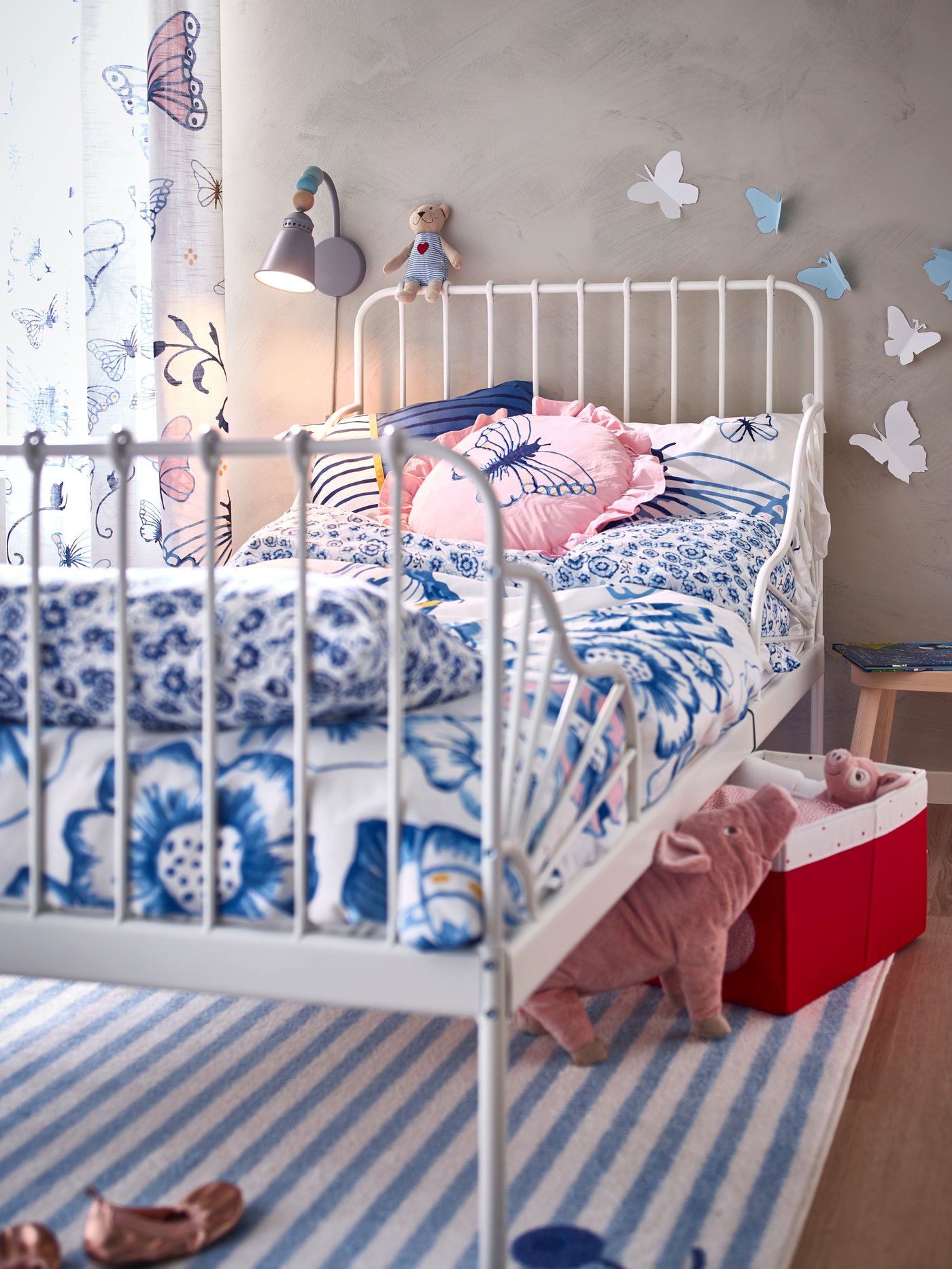 A MINNEN white extendable bed frame that has butterfly patterned bed linen on it, placed on a striped rug with soft toy pigs.