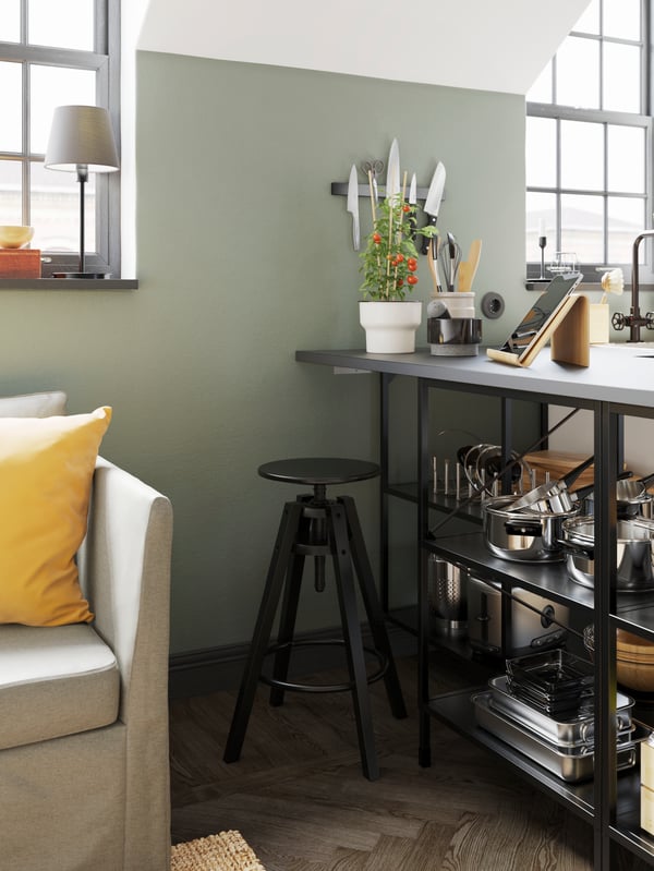 A black bar stool next to open shelves with a grey worktop holding cookware and a tablet stand and plant pot on top.