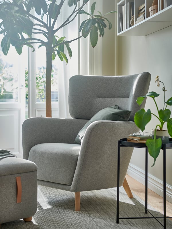 Partly sunlit through French doors, a beige-grey OSKARSHAMN wing chair and footstool stand by a large plant on a TIPHEDE rug.