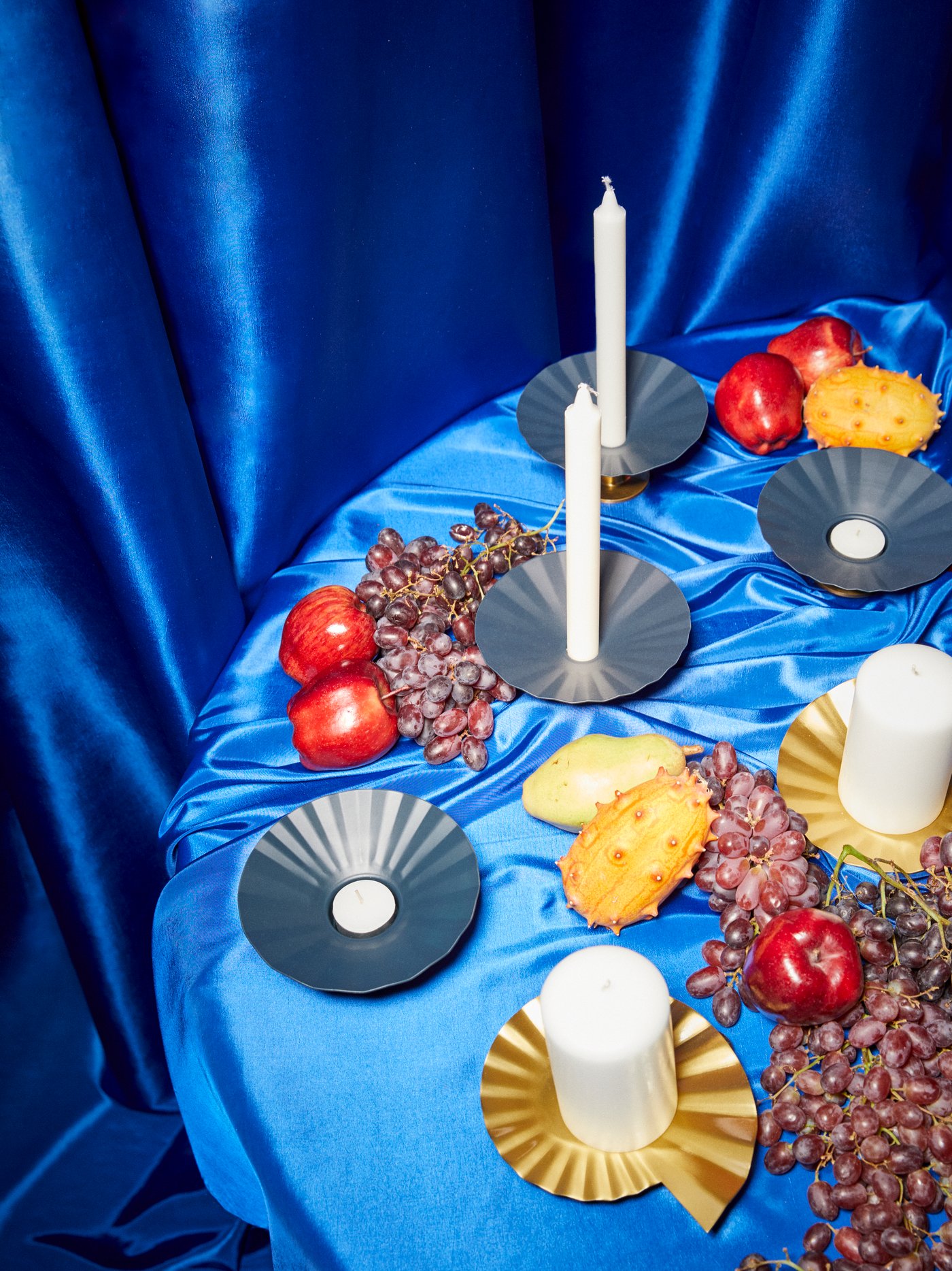 KARISMATISK metal candlesticks and tealight holders and gold candle dishes are on a table covered in a blue satin cloth.