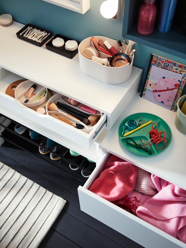 White shelf with drawers holding makeup products in storage boxes, pink clothing and trays with cotton pads and buds.