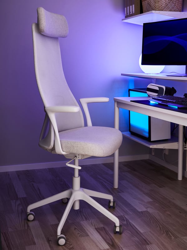 Una sedia da ufficio beige/bianca accanto a un tavolo e scaffali con un computer, uno schermo per computer e una lampada con luce blu.