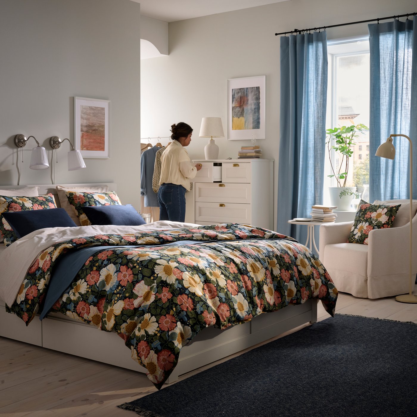 Es ist ein Schlafzimmer mit einem weißen FÅGELFJÄLLET Bettgestell mit Bettzeug in ARENBOK Bettwäsche mit Blumenmuster zu sehen. Eine Person räumt Kleidung in Schubladen ein.