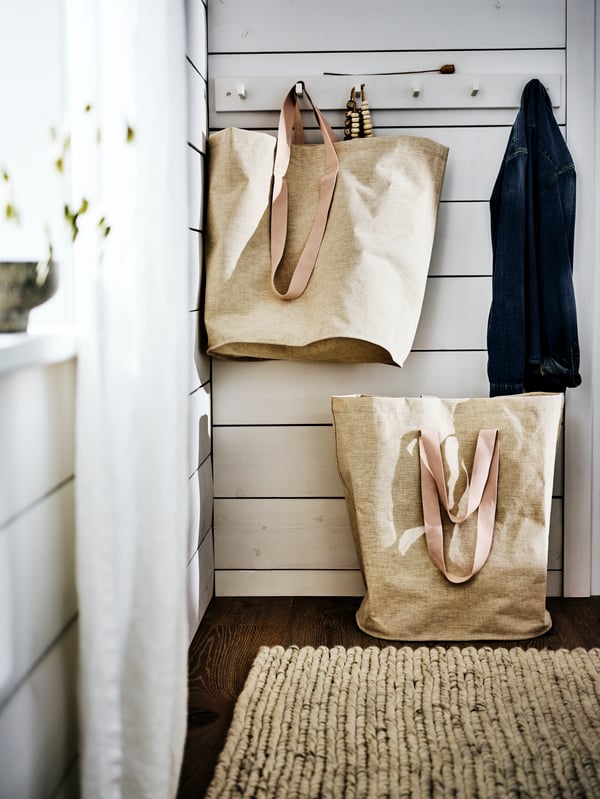 Un mur en lambris blanc avec des patères où un grand sac beige et un manteau sont accrochés. Un sac beige est par terre.